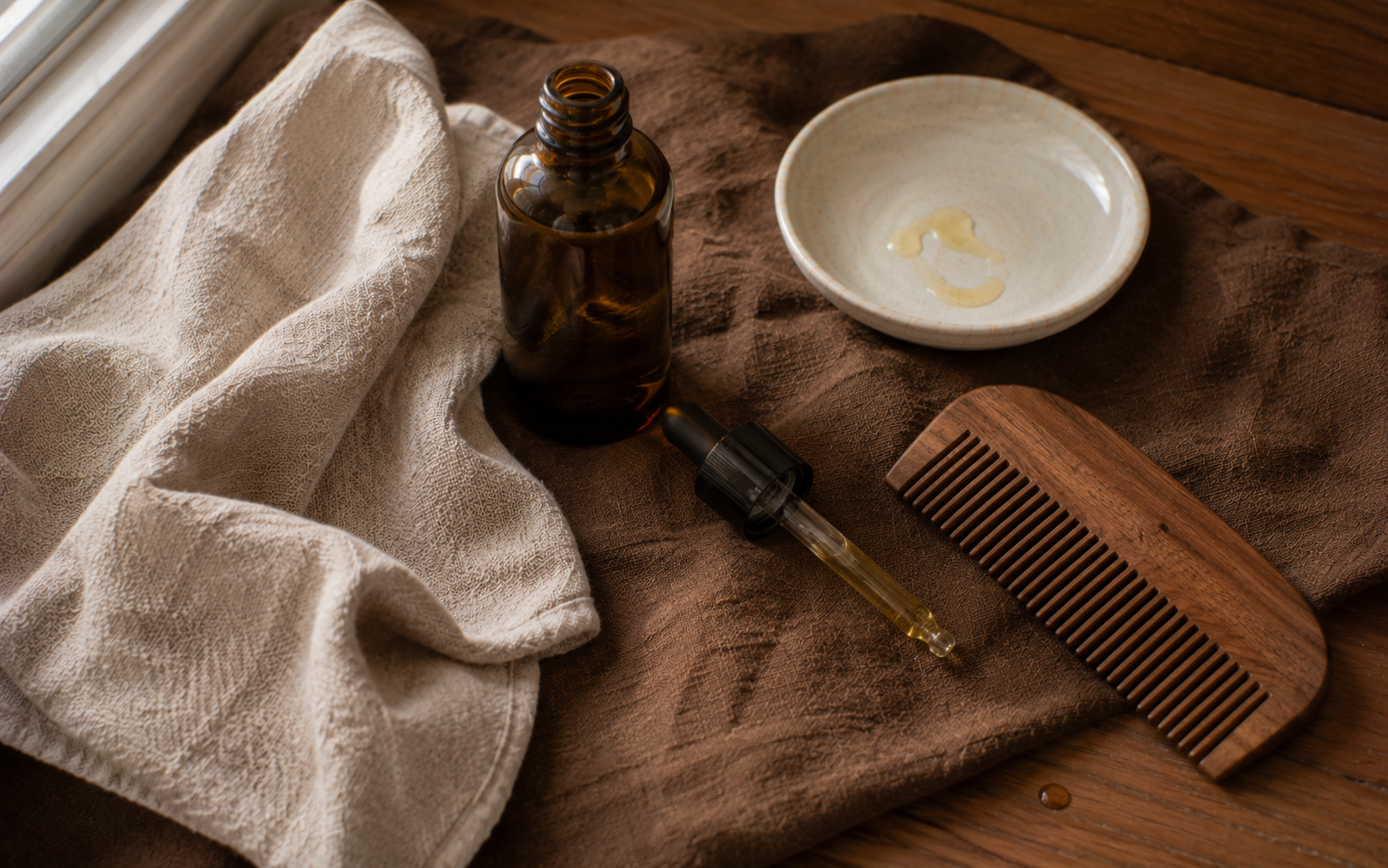 A small bottle of beard oil resting beside a folded oat milk-toned cloth and a wooden beard comb on rich tobacco brown linen in natural daylight