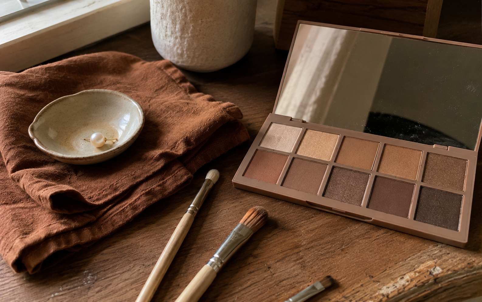 An open neutral eyeshadow palette in warm browns beside a few makeup brushes catching soft window light