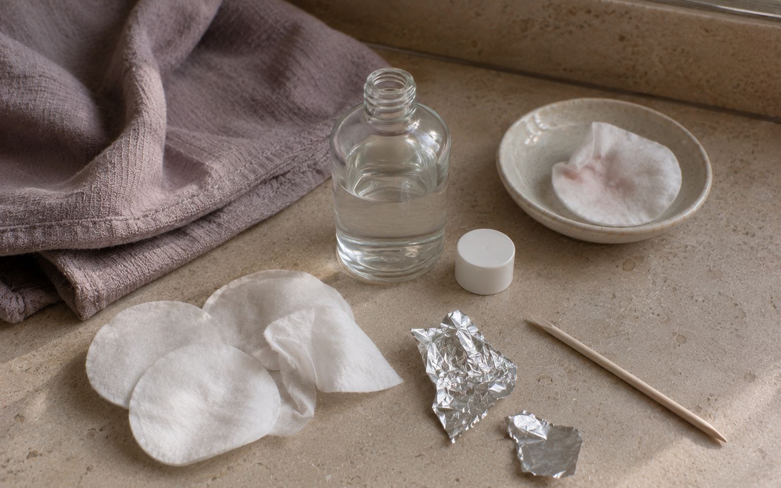 A small bottle of acetone, cotton pads, and aluminum foil pieces arranged on dusty mauve linen in natural light