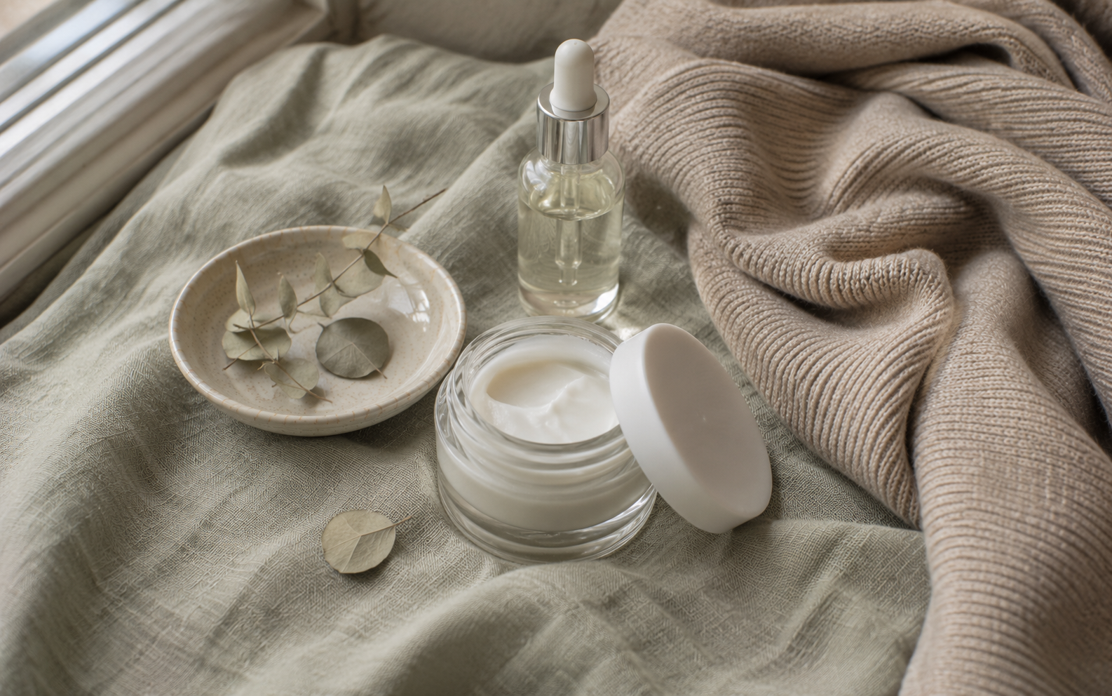 A small jar of rich face cream beside a folded warm cashmere scarf and dried eucalyptus on frosted sage linen in soft daylight